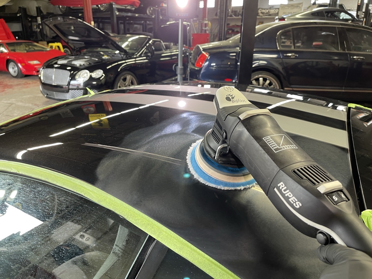 Rupes BigFoot dual-action polisher correcting paint on vehicle roof inside CBA Detailing professional shop in Dumont NJ with luxury vehicles in background